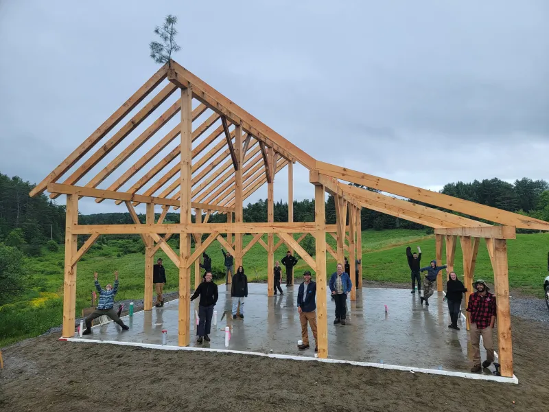 A finished timber frame stands against a grey sky with a whetting bush at the highest point. Students are posed in the structure, each standing by the post they made.