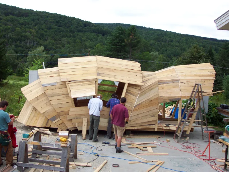 Builders constructing a faceted, armadillo-inspired wooden pavilion in an outdoor workshop set against a backdrop of green mountains.