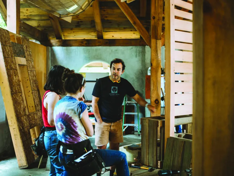 A man with dark curly hair and navy t-shirt shows a group of students a framed wall