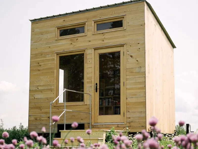 A finished tiny library stands in a field with sky in the background and red clover in the foreground.