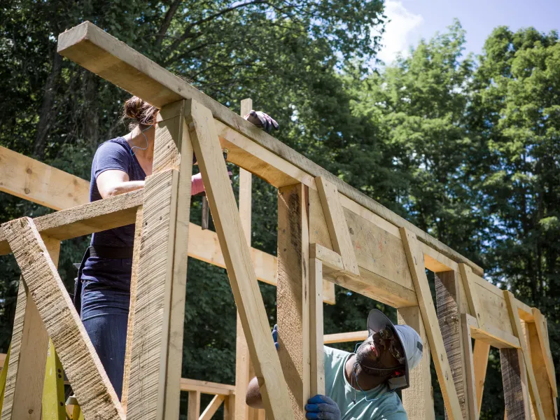 Two people work on framing a building with trees and sky in the background