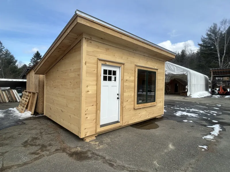 The exterior of a cabin with a large window, white door, pine shiplap siding and a shed roof.