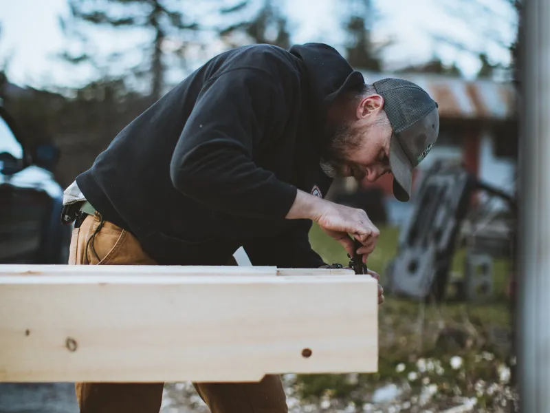 A student pictured outside on the pavement uses a combo square on a timber frame tenon.