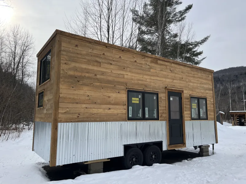 The exterior of a tiny house on wheels with wooden siding and a metal skirt.