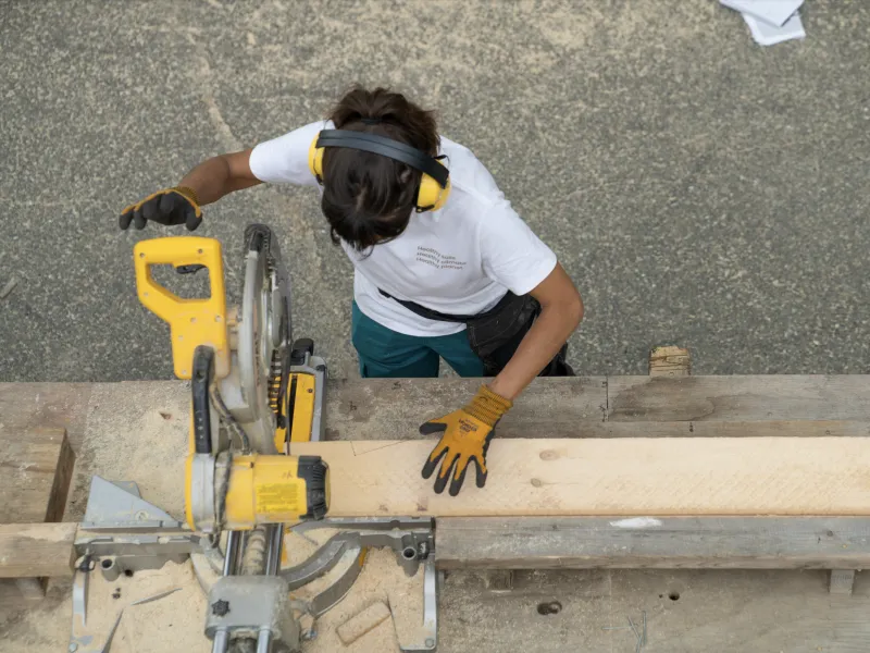 A student is photographed from above using a yellow chop saw to cut a piece of lumber. 