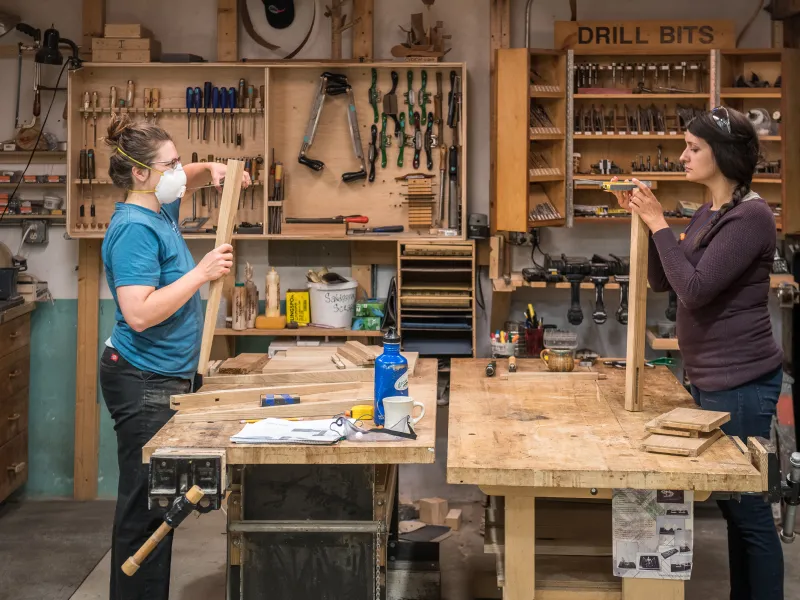 Two women at work benches facing each other measure pieces of wood in the woodshop