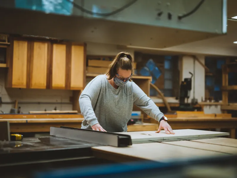 A person wearing a grey shirt and hair in a ponytail makes a cut on the table saw.
