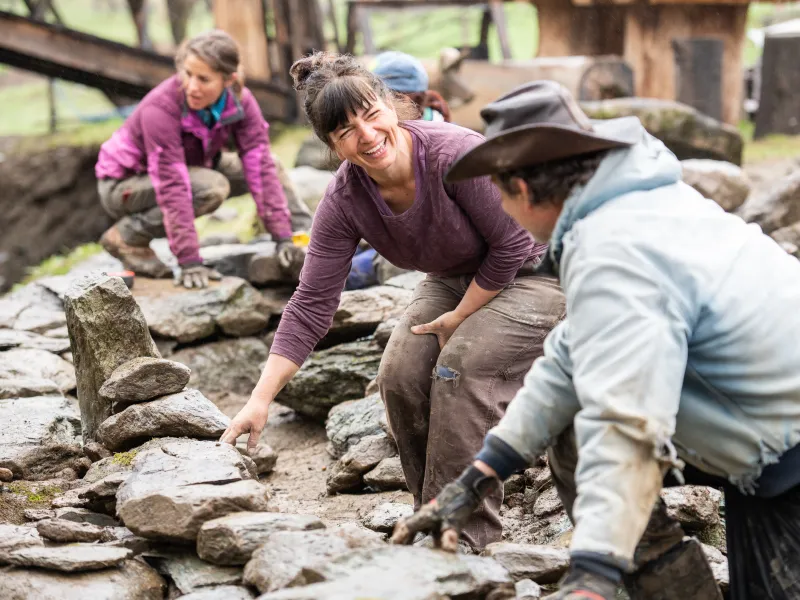A person with dark hair and purple shirt smiles at someone with their back to the camera. They are building a stone wall.