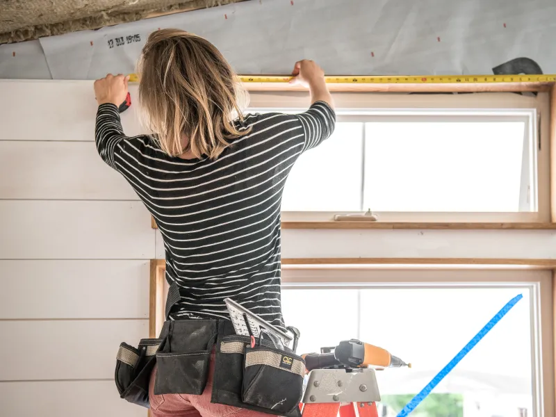A person with blonde hair and black-and-white striped shirt uses a measuring tape on the top of a door frame