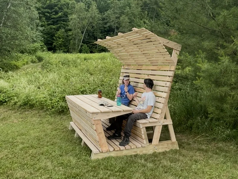 Two people sit on a picnic bench with an awning on a lawn