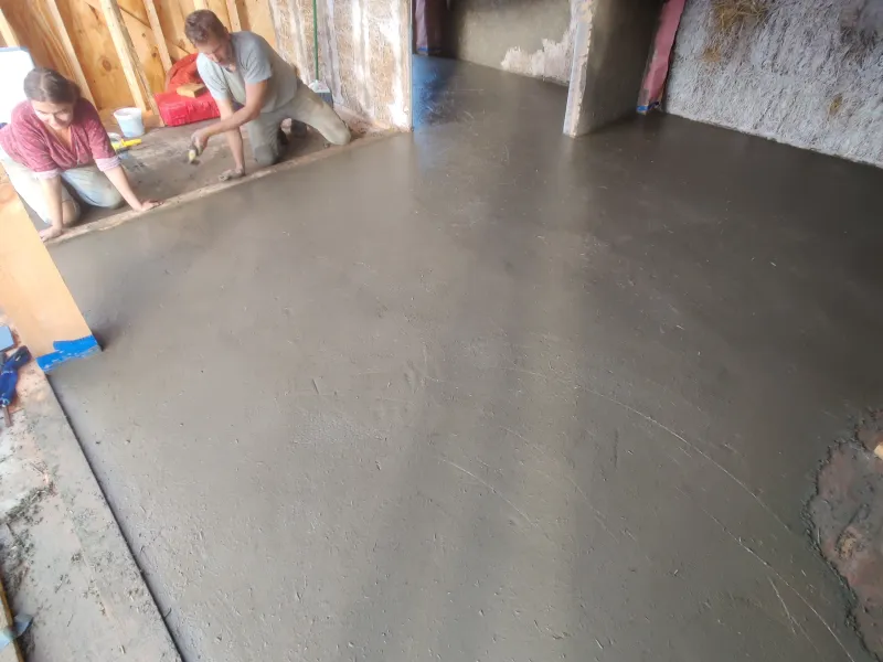An earthen floor inside a strawbale building with two people finishing the last edge details.