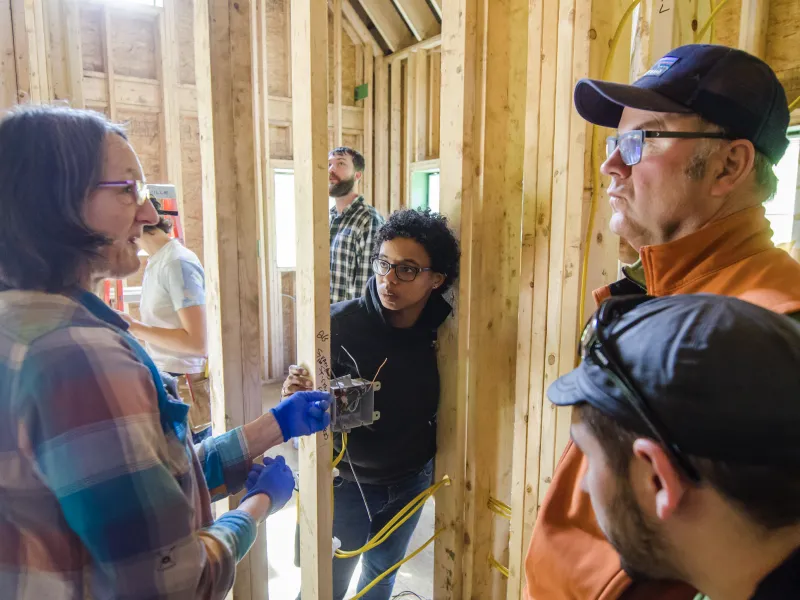 Students listen to an instructor gathered around a roughed-in electrical switch in a framed building.