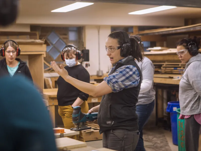 An instructor gestures in the woodshop in front of a group of students