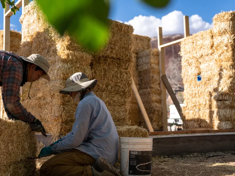 Two people crouch near a strawbale in front of a partially framed building.