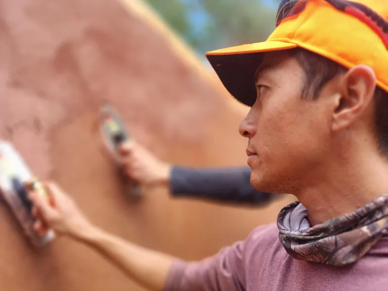 A person wearing an orange cap and neck gaiter carefully applies a smooth layer of reddish-brown clay plaster to a wall using a trowel. Another person’s hand is visible in the background, also plastering.