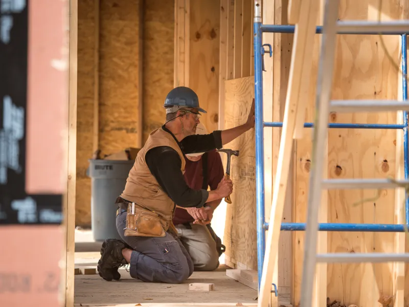 A person uses a hammer while kneeling inside a framed building with a hard hat on
