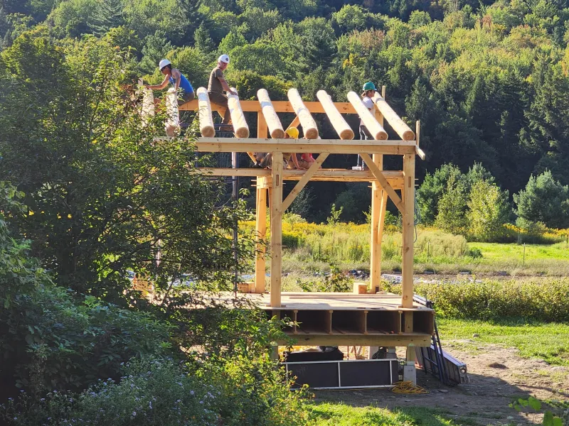 A group of people wearing safety helmets work together to construct a timber frame structure using round logs in a scenic outdoor setting. The partially built frame stands on a wooden platform surrounded by green grass, trees, and rolling forested hills under a clear blue sky.
