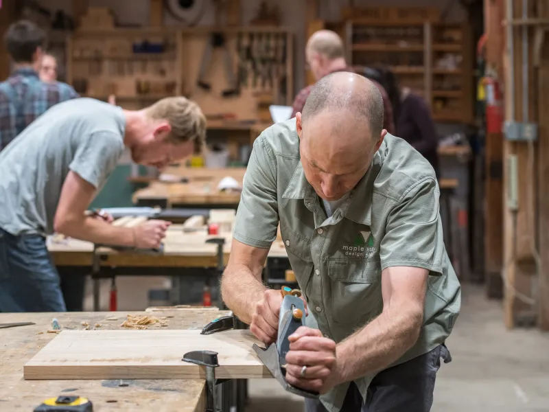 Two people, one in the foreground and one in the background, use hand planers in the woodshop.