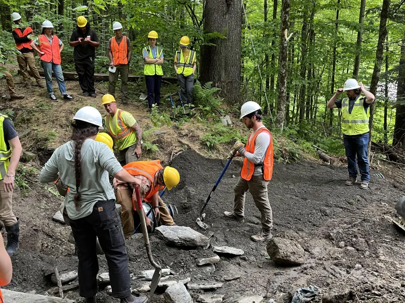 Gemini said A group of workers and students wearing safety vests and hard hats collaborate on a culvert installation project, using hand tools to move large stones and dark soil in a lush, wooded environment.