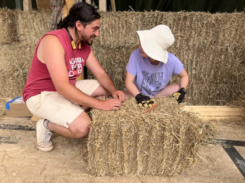 Two people work together shaping a straw bale inside a partially built structure made of straw walls. One person wears a sleeveless red shirt and light shorts, while the other wears a wide-brimmed hat, gloves, and a purple shirt.