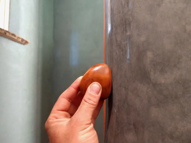 A close up shot of a hand using a red stone to burnish a gray plastered wall.
