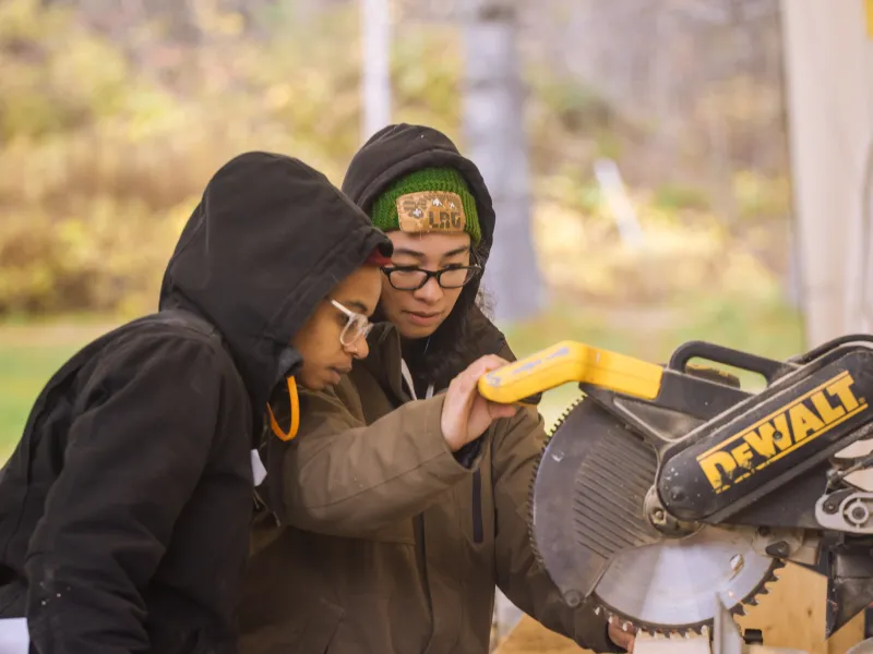 An instructor guides a student at a chop saw station in an outdoor classroom.