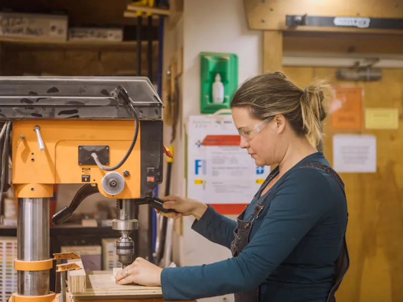 A student with blonde hair in a ponytail wearing a turquoise shirt uses the drill press in the woodshop.