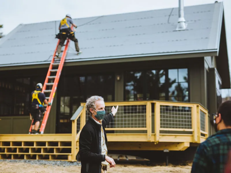 A man in the foreground gestures at a house in the background. Workers are installing solar panels on the roof.