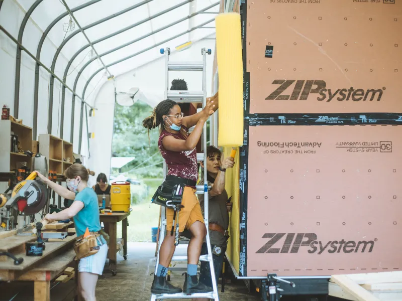 Students work on a tiny house in an outdoor classroom. A student with braids uses a nail gun on the tiny house siding in the foreground.