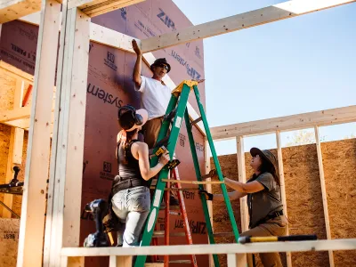An instructor on a ladder gestures at a partially framed building. 