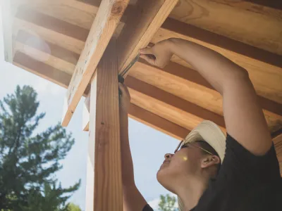 A person uses an angle finder on the corner of an outdoor structure, with trees and sky in the background. The photo is shot from below, looking up at the person reaching up to the corner. 