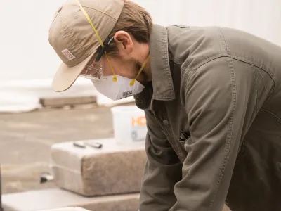 A student wearing a hat and dust mask sccops hemp out of a wheelbarrow