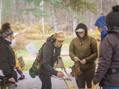 Several people standing outside look at a piece of lumber. The teacher is pointing at the lumber to show the students measurements.