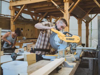 A person with buzzed hair uses a chop saw to cut lumber in a woodshop.