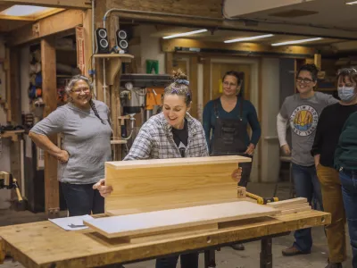 An instructor holds a piece of wood on a work bench, demonstrating for students standing behind.