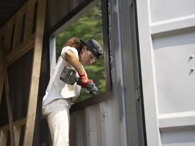A person in white clothing uses a driver on the window of a shipping container building.