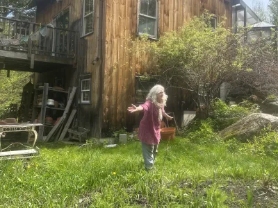 Heidi stands in her lawn in front of her house, with her arms wide. She has long gray hair and a pink top. 