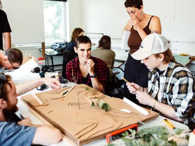 A group of students gather around a topographic cardboard model in a studio classroom.