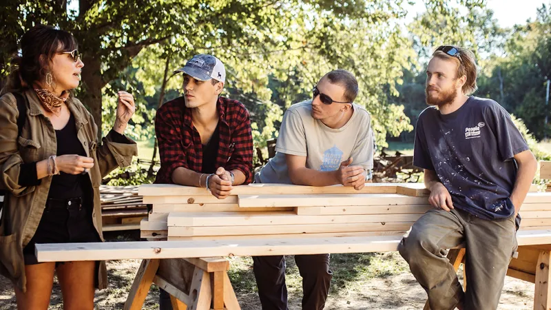 An instructor speaks to a group of students leaning against a pile of lumber on sawhorses.