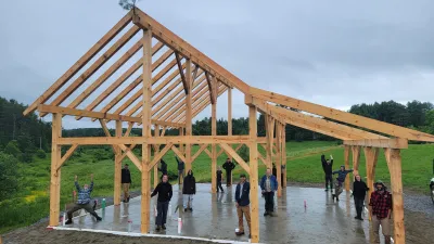 A finished timber frame stands against a grey sky with a whetting bush at the highest point. Students are posed in the structure, each standing by the post they made.