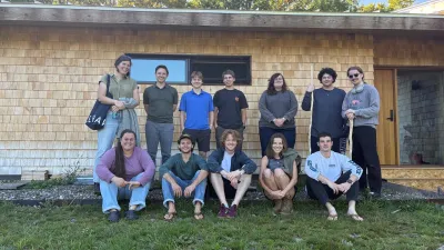 The 2026 Design/Build semester poses for a group photo outside in front of a shingled cabin. There are two rows of students, some sitting down in front. It is a sunny day with blue skies.