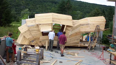 Builders constructing a faceted, armadillo-inspired wooden pavilion in an outdoor workshop set against a backdrop of green mountains.