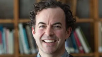 Britton Rogers, standing in front of books, smiles at the camera. He has short, dark curly hair and a blue sweater.