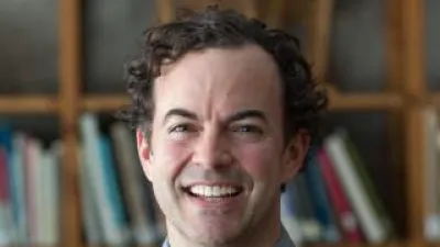 Britton Rogers, standing in front of books, smiles at the camera. He has short, dark curly hair and a blue sweater.