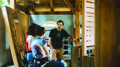 A man with dark curly hair and navy t-shirt shows a group of students a framed wall