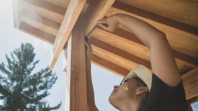 A person uses an angle finder on the corner of an outdoor structure, with trees and sky in the background. The photo is shot from below, looking up at the person reaching up to the corner. 