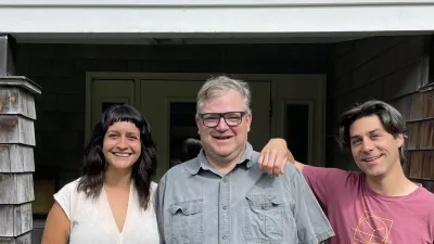 Three people stand smiling in front of the Yestermorrow front entrance sign. Left to right they are a shorter, dark haired femme person with a beige shirt and black pants, a man with a blue-gray light blue button down and glasses, and a man with longer, dark hair wearing a pink yestermorrow t-shirt