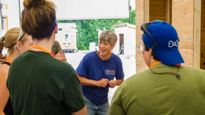 Patti is smiling, talking to a group of students with their backs to the camera. Patti has short gray hair and is wearing a blue t-shirt.