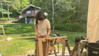 A semester student with long dark hair measures a piece of lumber on sawhorses.