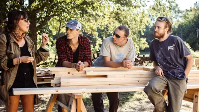 An instructor speaks to a group of students leaning against a pile of lumber on sawhorses.